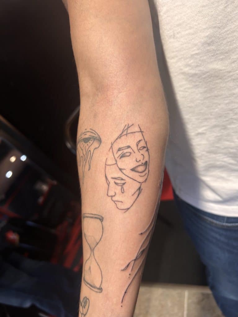 Fine line blackwork tattoo on a person's forearm featuring classic comedy and tragedy drama masks