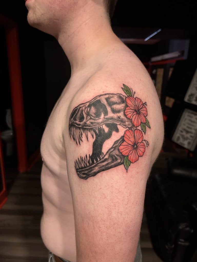 Black and grey tattoo of a T-Rex skull surrounded by vibrant pinkish-red hibiscus flowers on a man's upper arm.