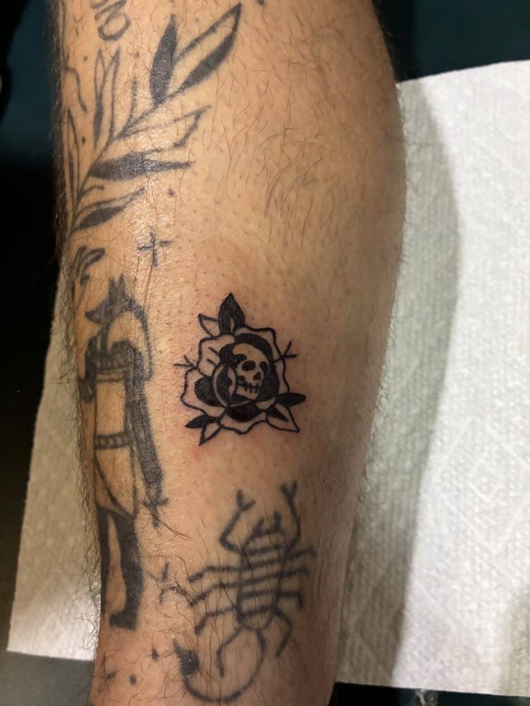 A classic black and grey traditional style tattoo of a skull and rose on a lower leg, custom designed at Mayhem Ink Tattoo in Nashua, NH.